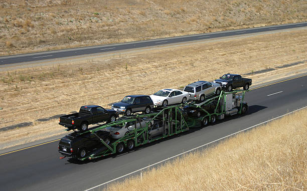 A green car carrier truck from AZ Auto Transport Glendale transports several vehicles, including SUVs and sedans, along a highway bordered by dry grass and dirt.