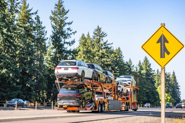 A car carrier truck transports multiple vehicles on a highway lined with trees. A yellow merge traffic sign is visible on the right side of the road, illustrating a typical scene encountered during AZ Car Shipping Glendale services.